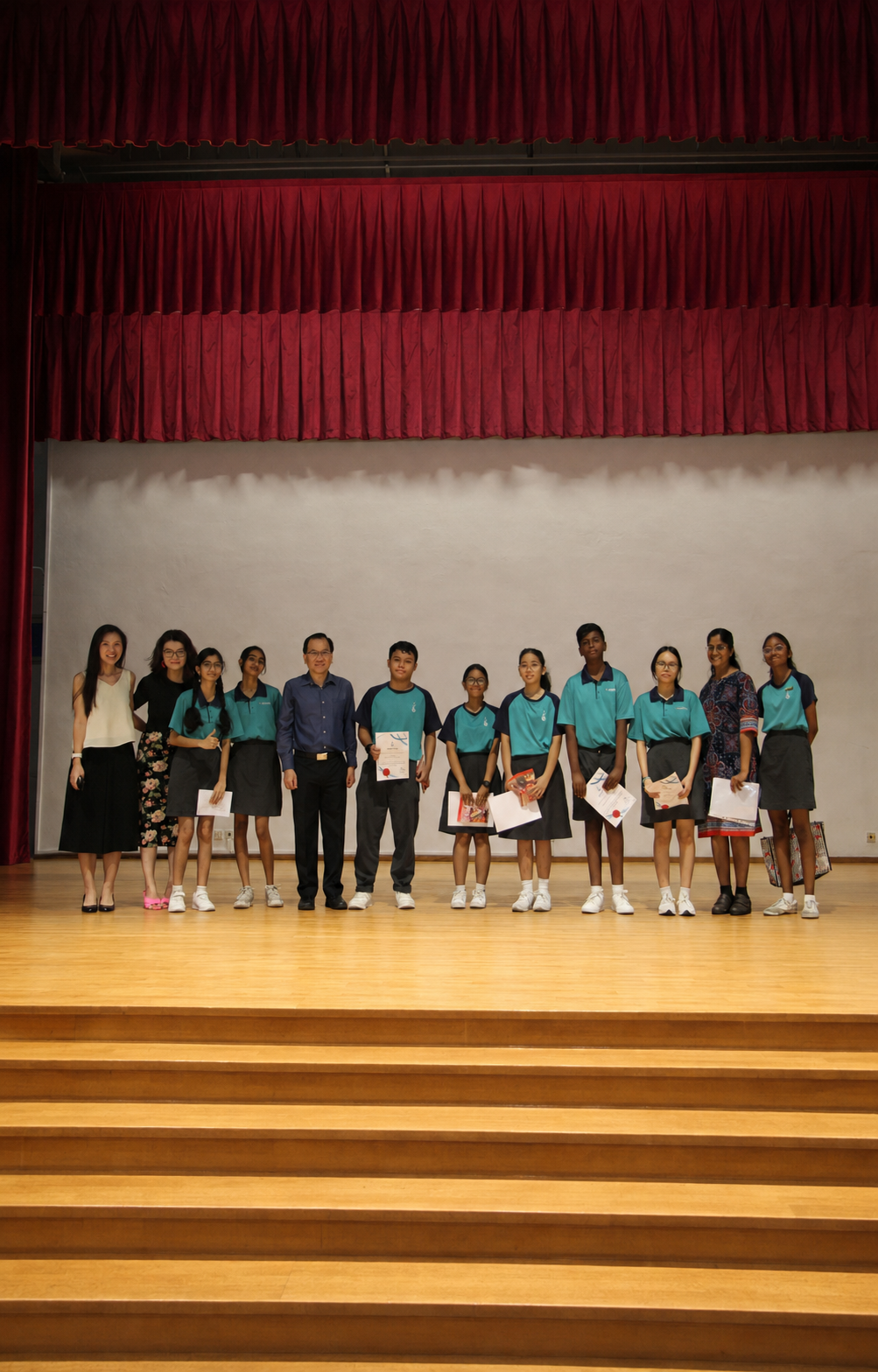 A group of adults and students pose on stage with prizes